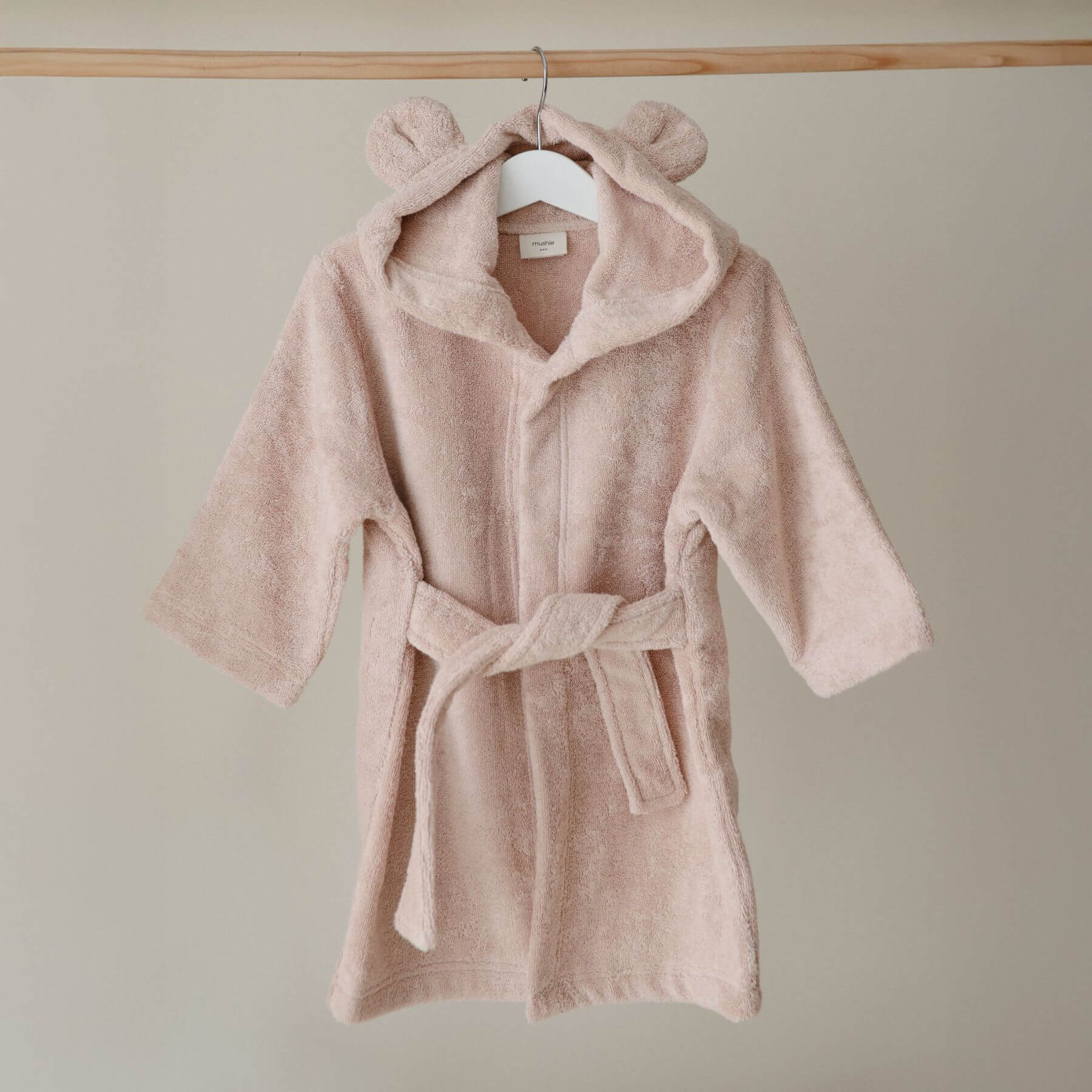 Mushie Bear Robe on a hanger against a beige wall.