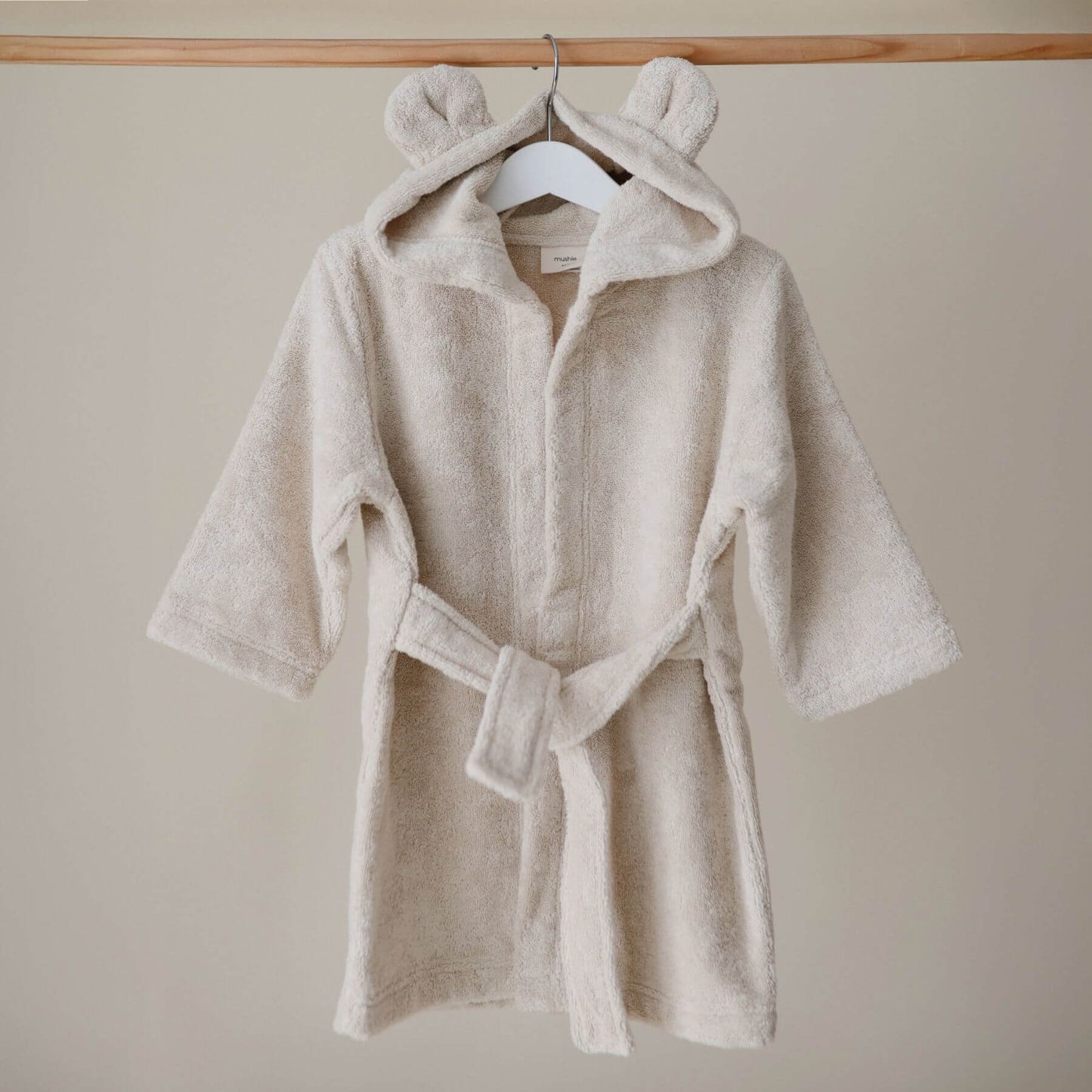 Mushie Bear Robe Beige on a hanger against a beige wall
