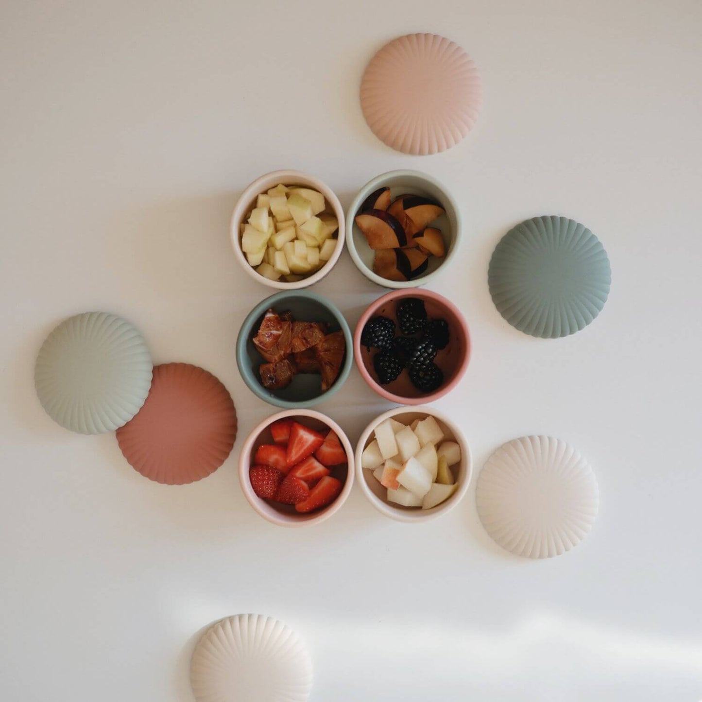 Mushie Silicone Baby Food Containers, each filled with different foods, alongside six matching lid-like pieces.