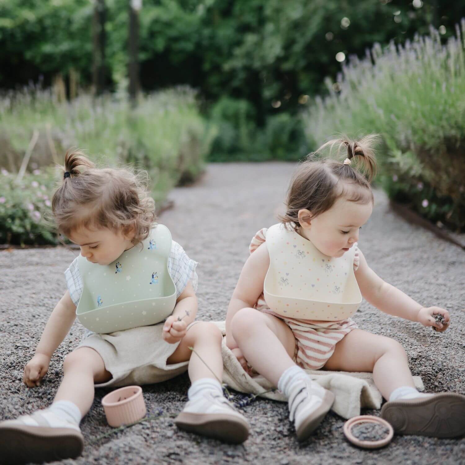 Two young children sitting on a path outdoors, wearing bibs.