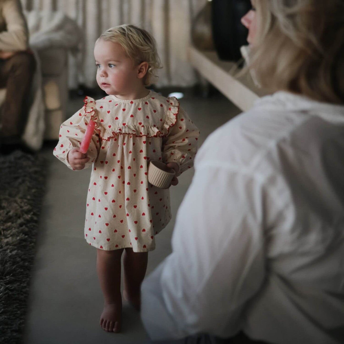 Young girl in a polka dot dress holding a food container, with an adult in a white shirt in the foreground.