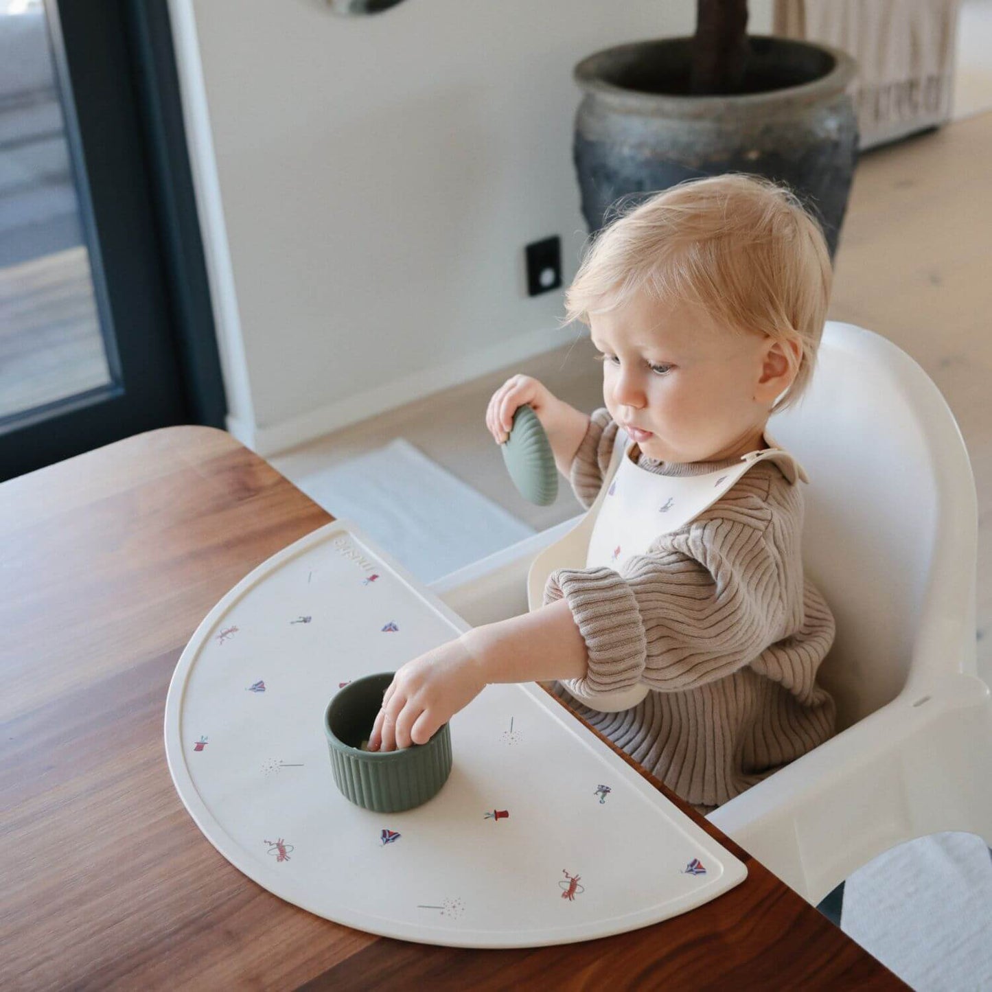 Child in a high chair with a placemat, eating from a green food container