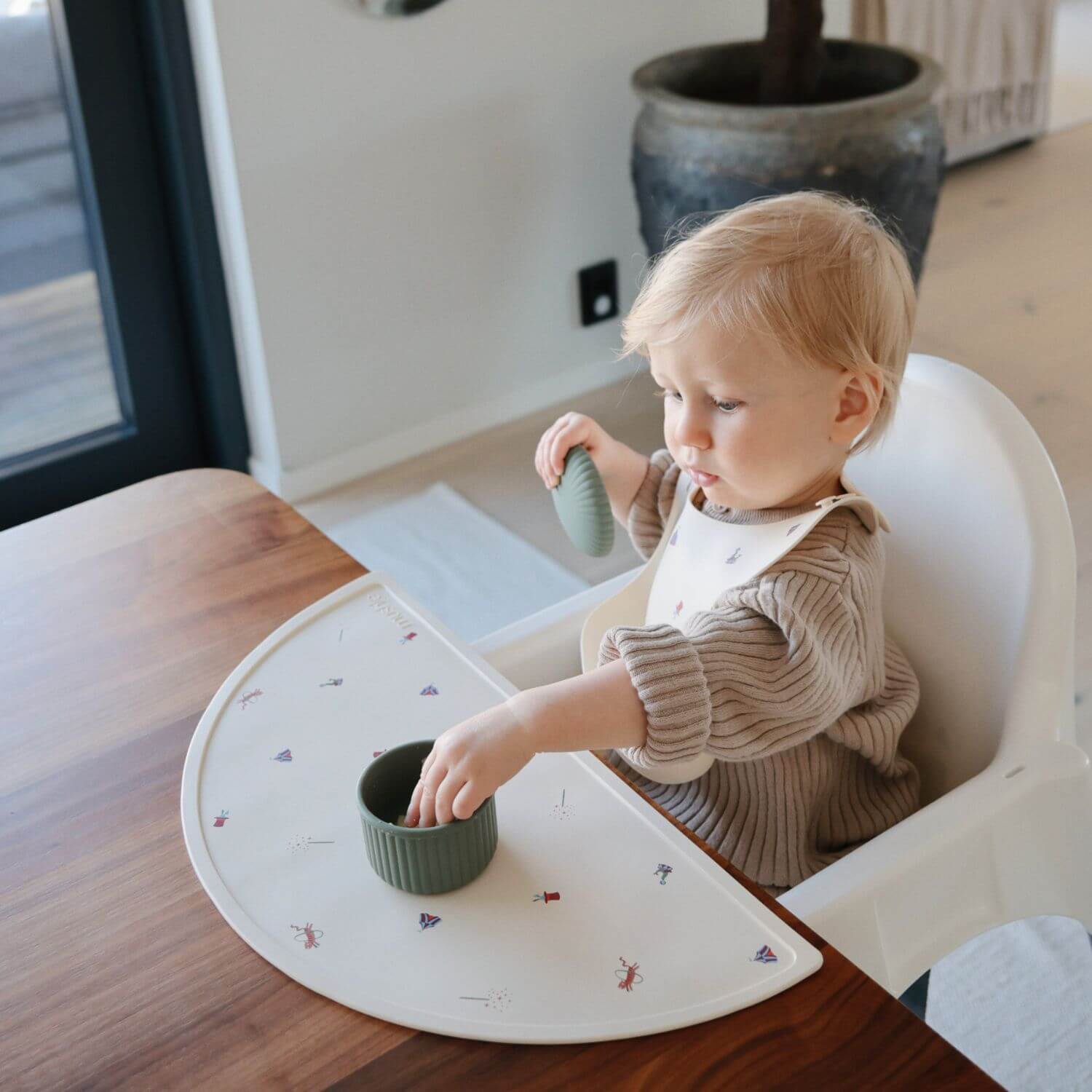 Child in a high chair with a placemat, eating from a green food container