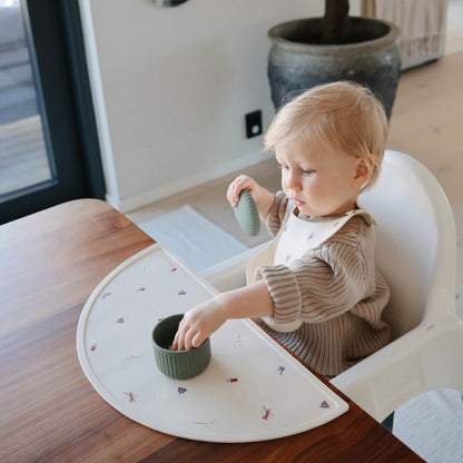 Child in a high chair with a placemat, eating from a green food container
