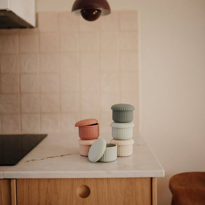 Mushie Silicone Baby Food Containers in muted pastel colors arranged on a kitchen countertop