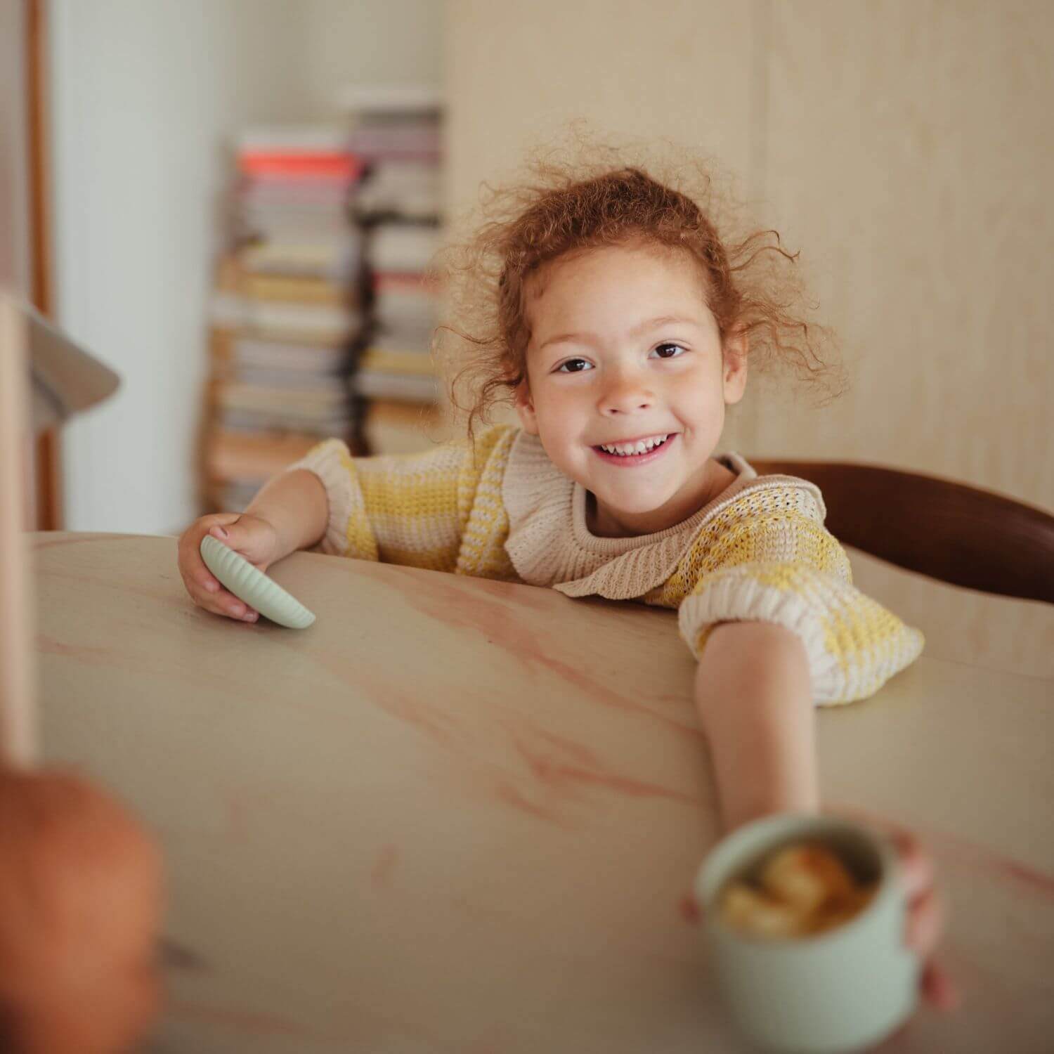 A young child holds a Mushie Silicone Baby Food Container in one hand and a small round object in the other.