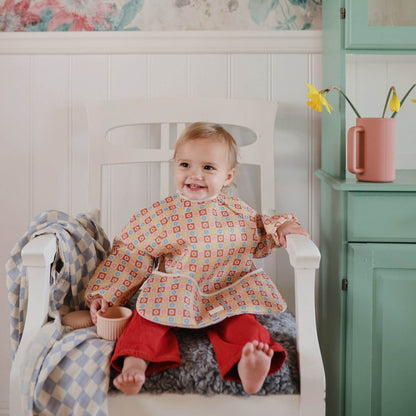 Baby sitting in a chair wearing a patterned bib and red pants, with a floral wall and green cabinet in the background.