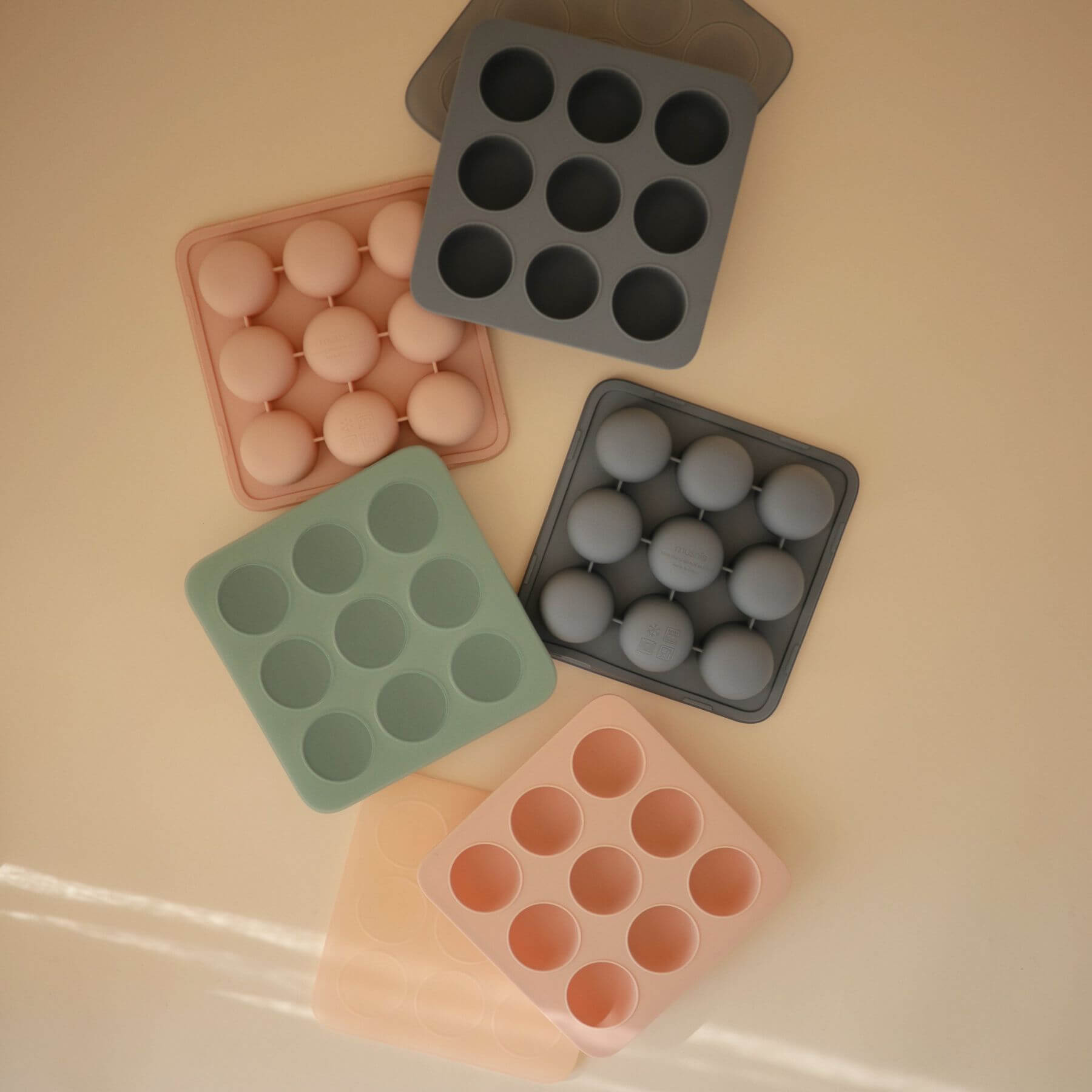 Collection of Mushie Baby Food Freezer Trays in various colors on a beige surface