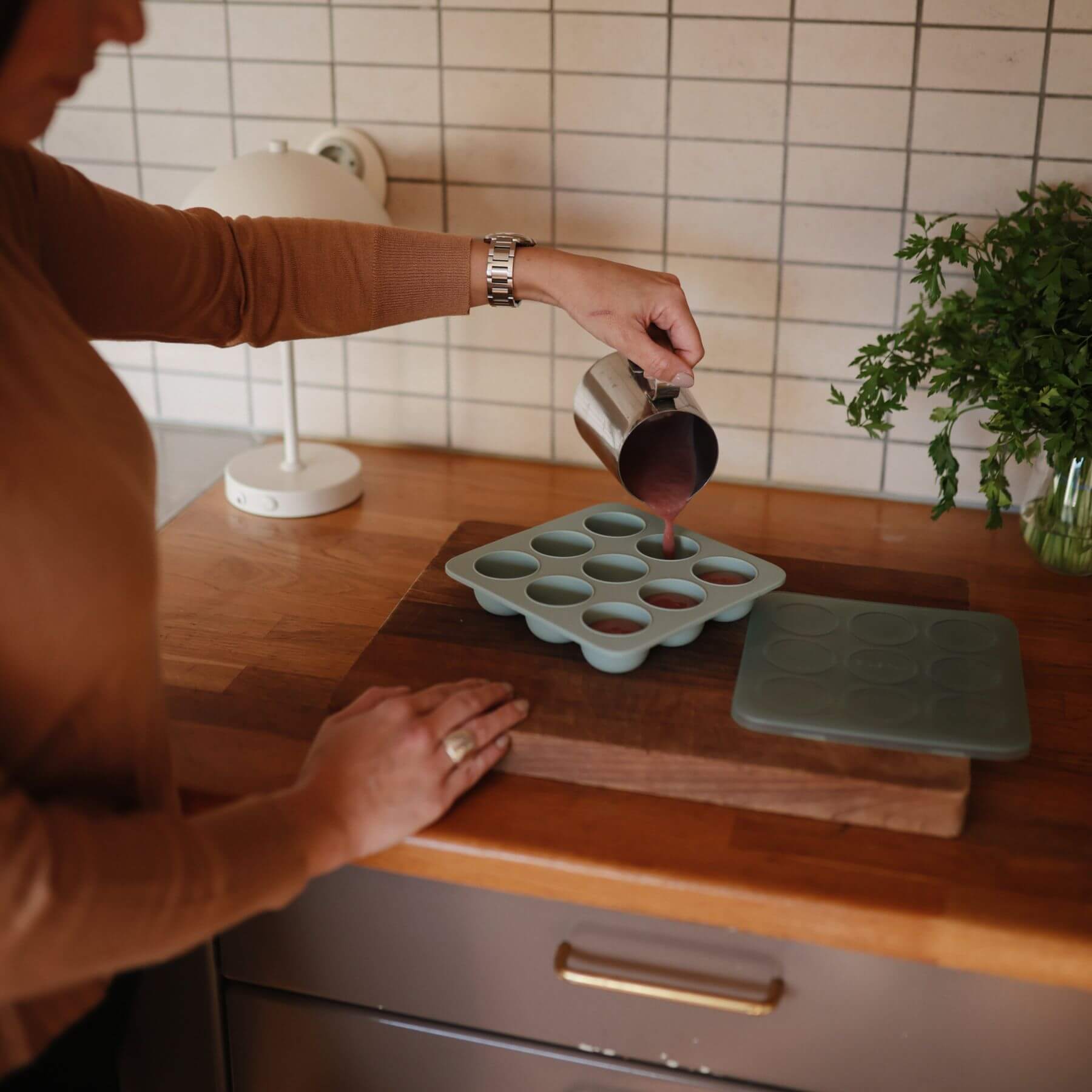 Person pouring liquid into a Mushie Baby Food Freezer Tray Cambridge Blue on a wooden cutting board in a kitchen.