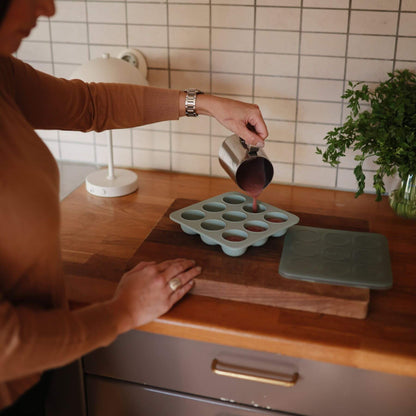 Person pouring liquid into a Mushie Baby Food Freezer Tray Cambridge Blue on a wooden cutting board in a kitchen.