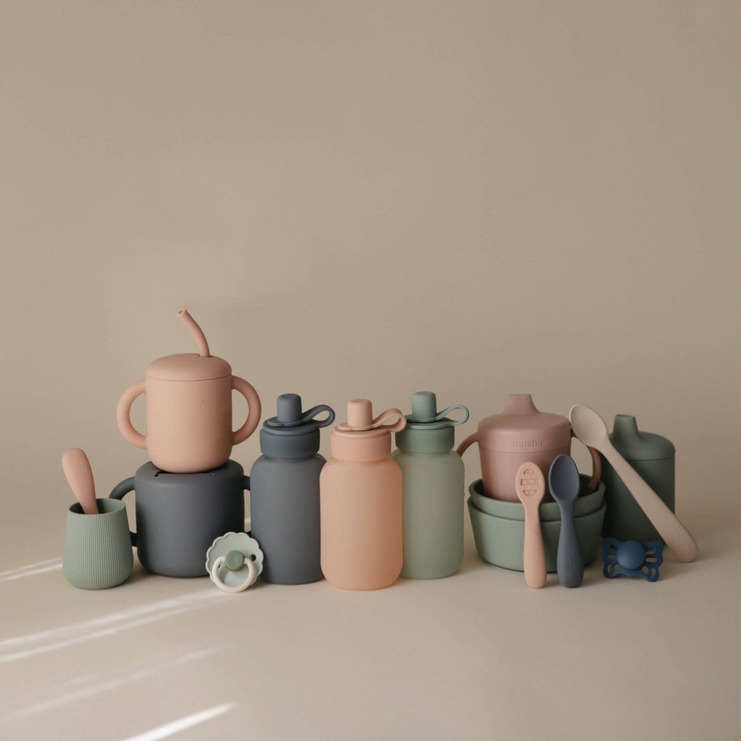 Collection of colorful children's cups, bottles, and utensils on a beige surface with a neutral background.