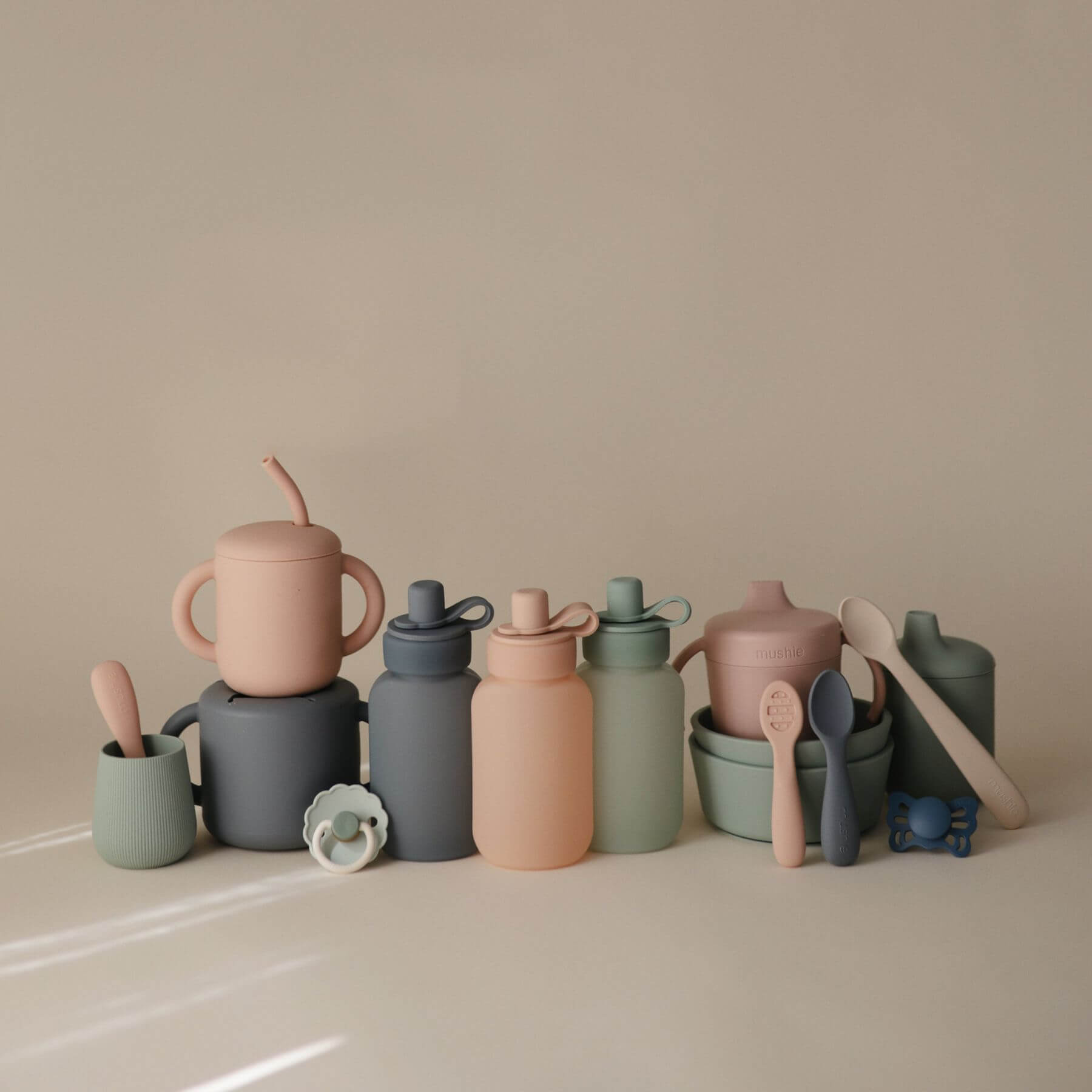 Collection of colorful children's cups, bottles, and utensils on a beige surface with a neutral background.