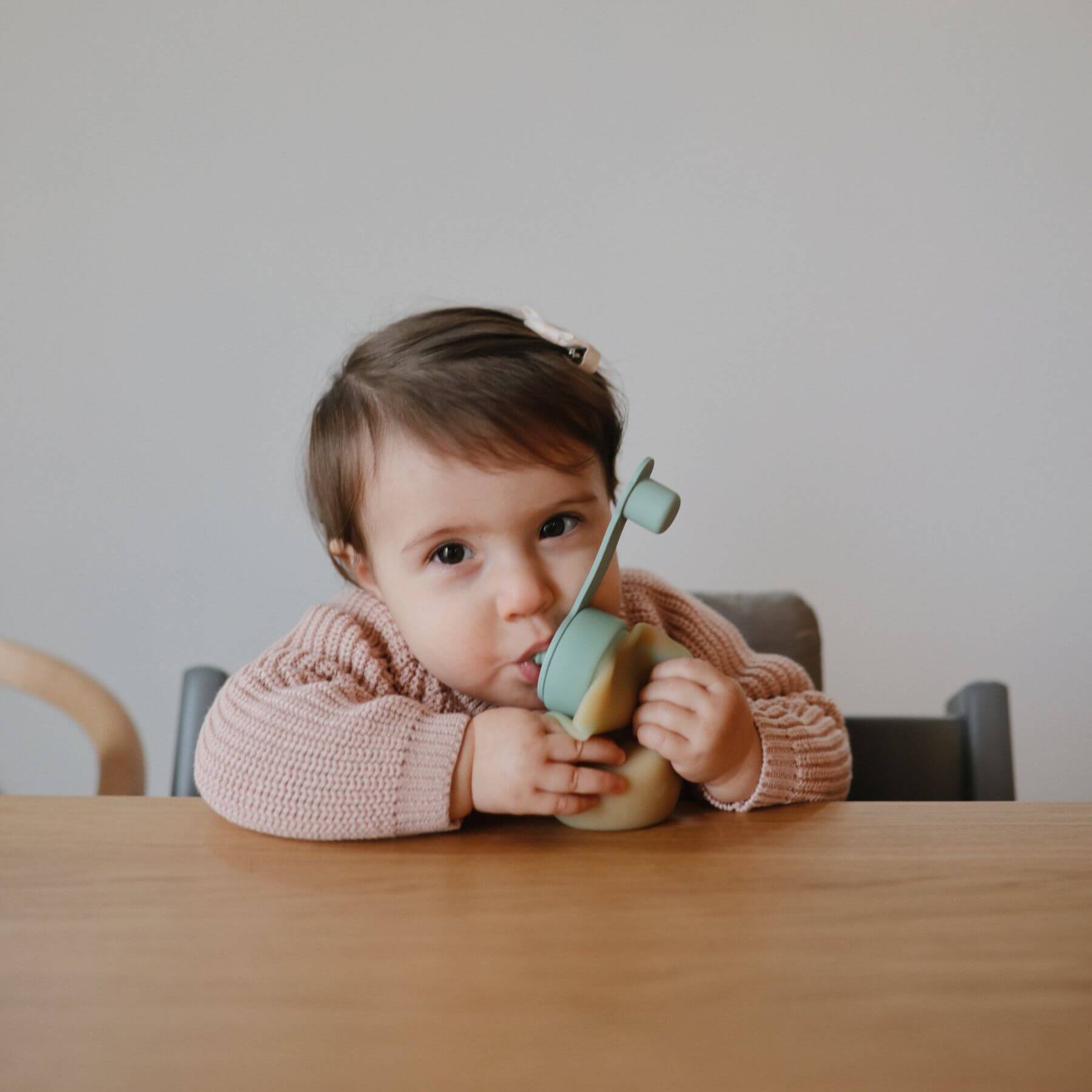 Child sitting at a table holding a Mushie Silicone Puree Pouch, wearing a pink sweater.
