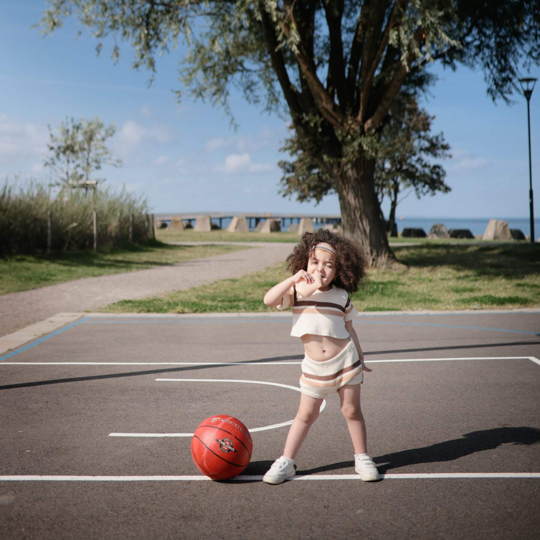 Child playing with a basketball on an outdoor court with trees and a path in the background