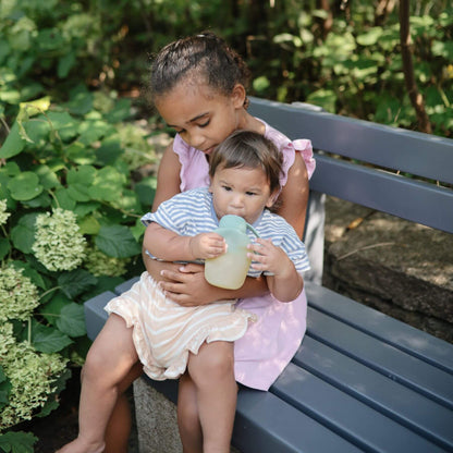 Two young girls sitting on a bench outdoors, one holding a sippy cup.