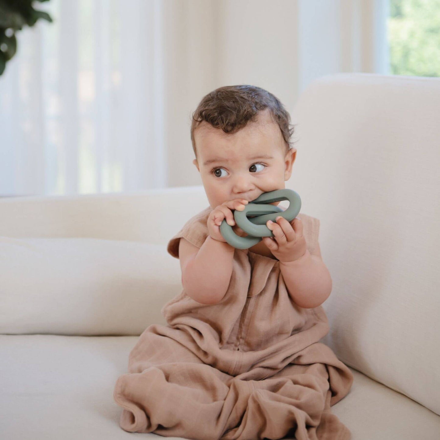 Baby in Mushie Organic Cotton Sleep Bag, sitting on a couch holding a green teething ring