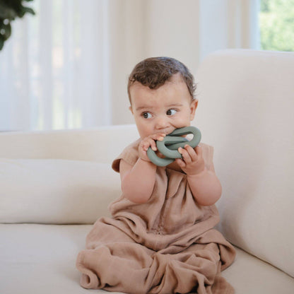 Baby in Mushie Organic Cotton Sleep Bag, sitting on a couch holding a green teething ring