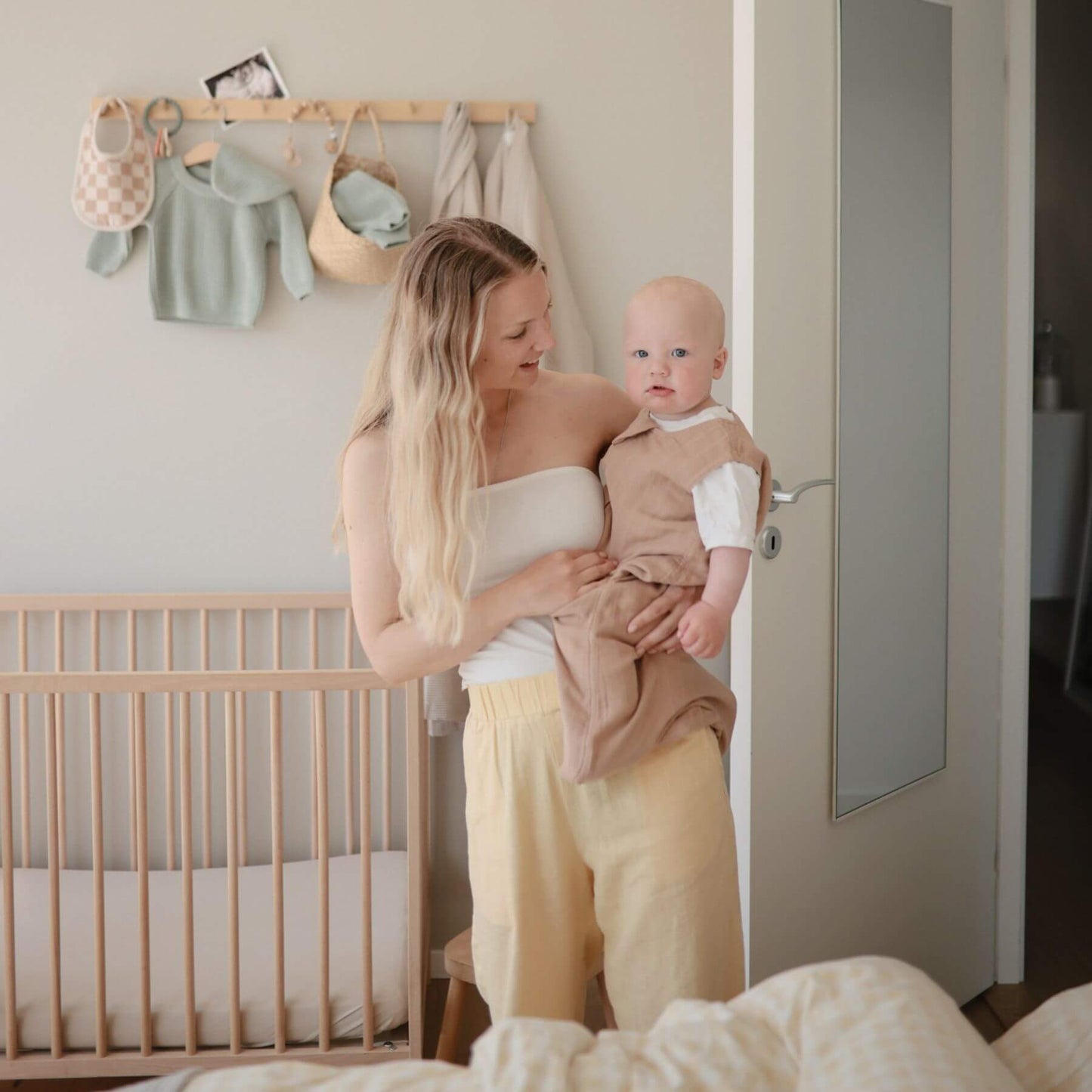 Woman holding a baby in a nursery with a crib and hanging clothes in the background.