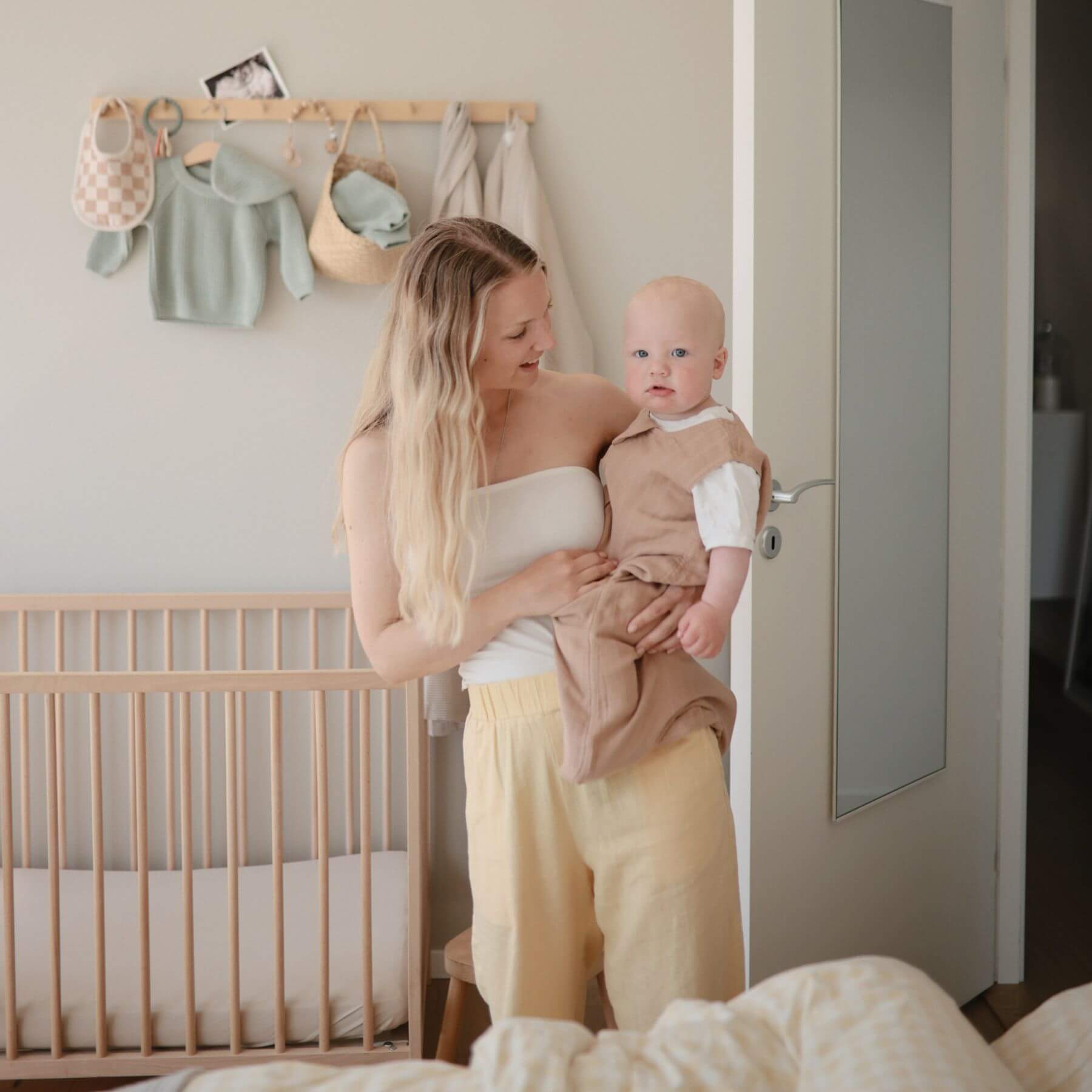 Woman holding a baby in a nursery with a crib and hanging clothes in the background.