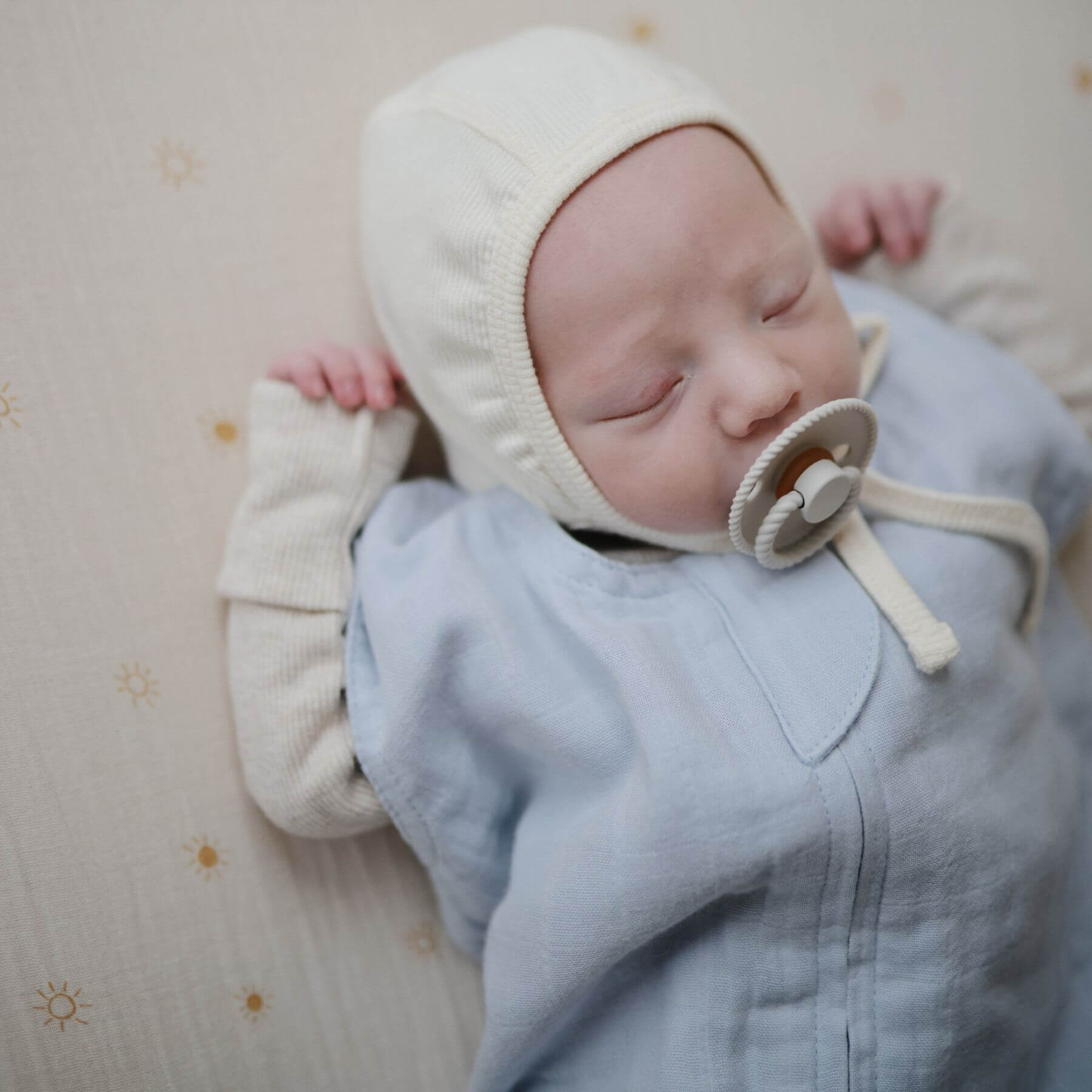 Newborn baby wearing Mushie Organic Cotton Sleep Bag and cream bonnet with a pacifier, lying on a soft surface.