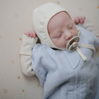Newborn baby wearing Mushie Organic Cotton Sleep Bag and cream bonnet with a pacifier, lying on a soft surface.