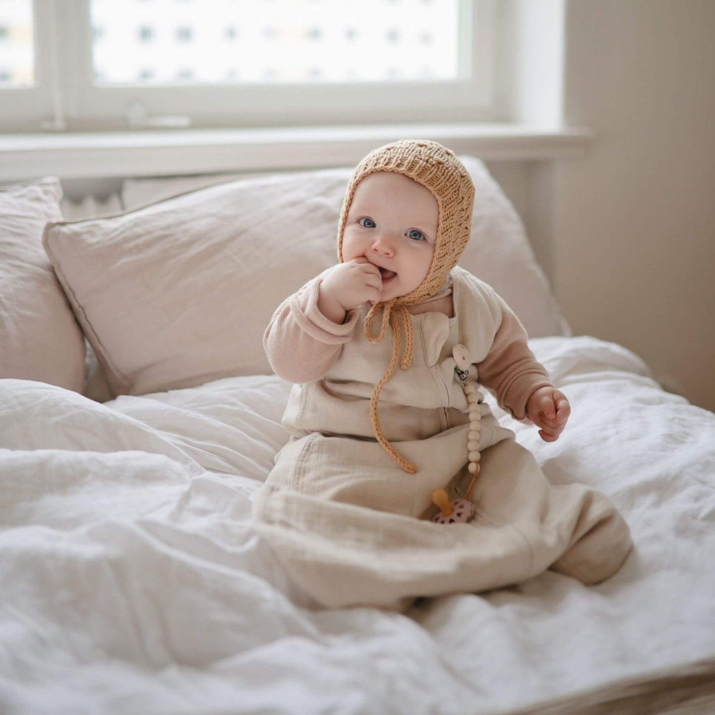 Baby sitting on a bed wearing a beige bonnet and Mushie Organic Cotton Sleep Bag, with a soft focus background.