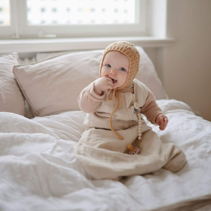 Baby sitting on a bed wearing a beige bonnet and Mushie Organic Cotton Sleep Bag, with a soft focus background.