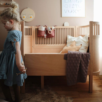 Child in a blue dress standing next to a wooden crib with a teddy bear and toys.