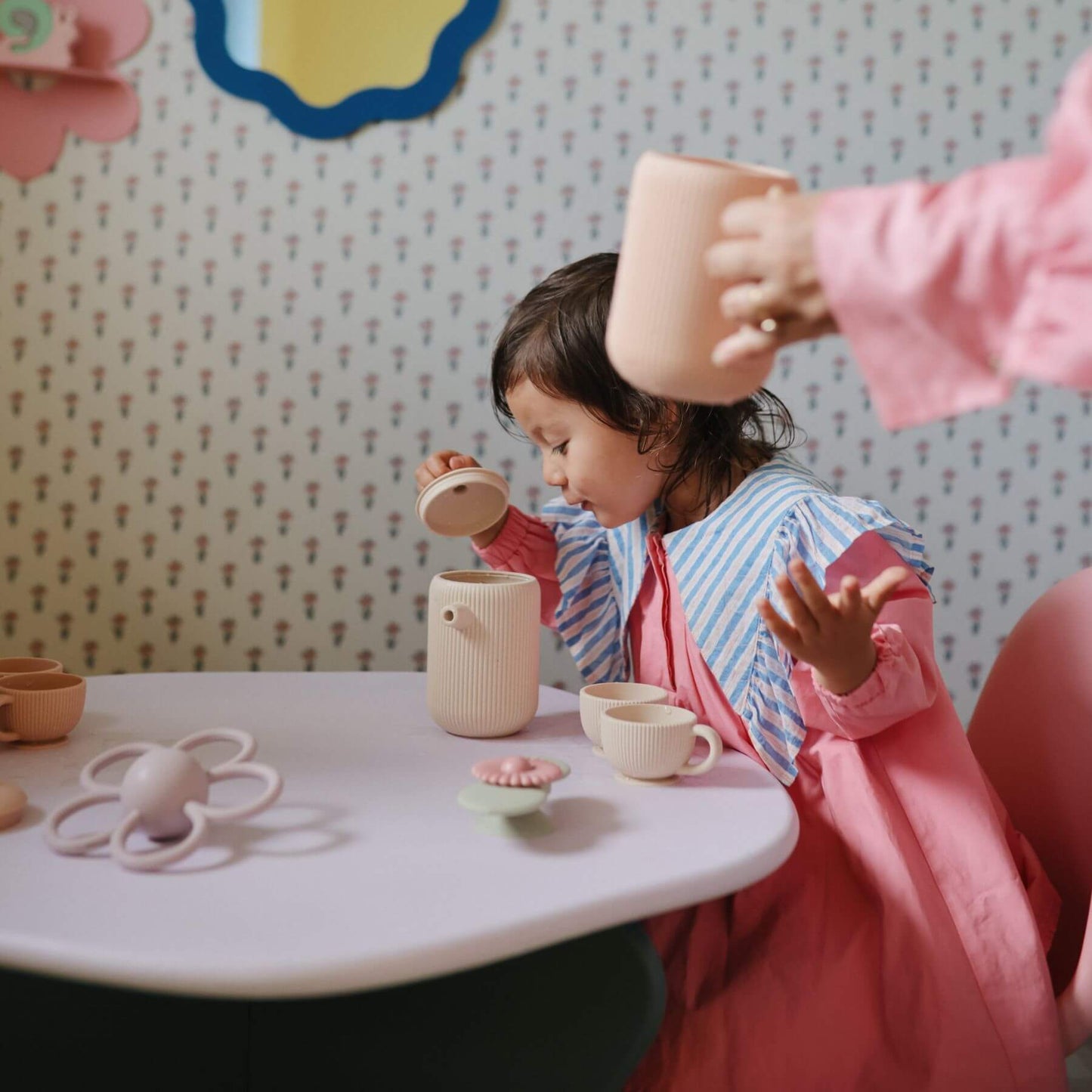 Child playing with Mushie Silicone Tea Play Set at a table against a patterned wall.