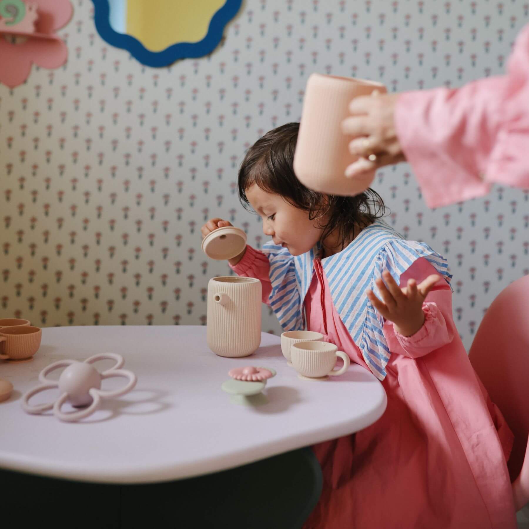Child playing with Mushie Silicone Tea Play Set at a table against a patterned wall.