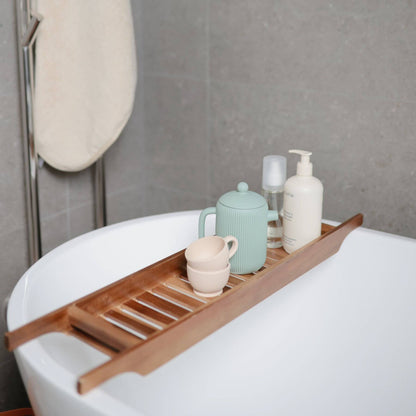 Wooden bathtub tray with bathroom items on a white bathtub against a gray tiled wall.