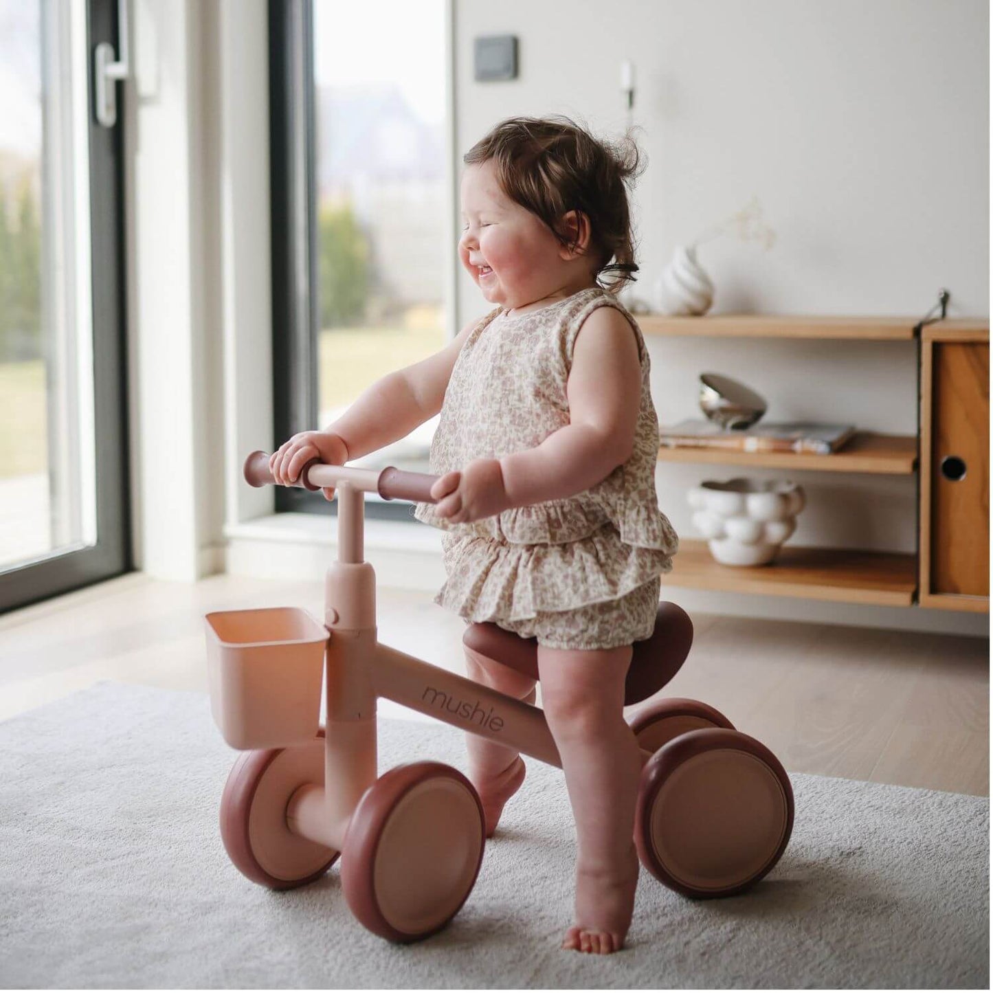 A toddler stands on Mushie Mini Toddler Bike, indoors near a window and wooden shelf.