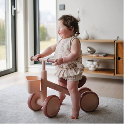 A toddler stands on Mushie Mini Toddler Bike, indoors near a window and wooden shelf.