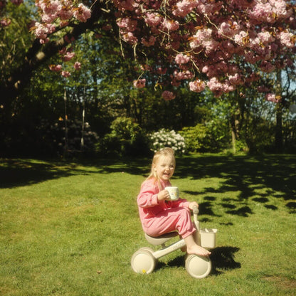 A young child sitting on Mushie Mini Toddler Bike in a grassy backyard under a flowering cherry tree, holding a white mug.