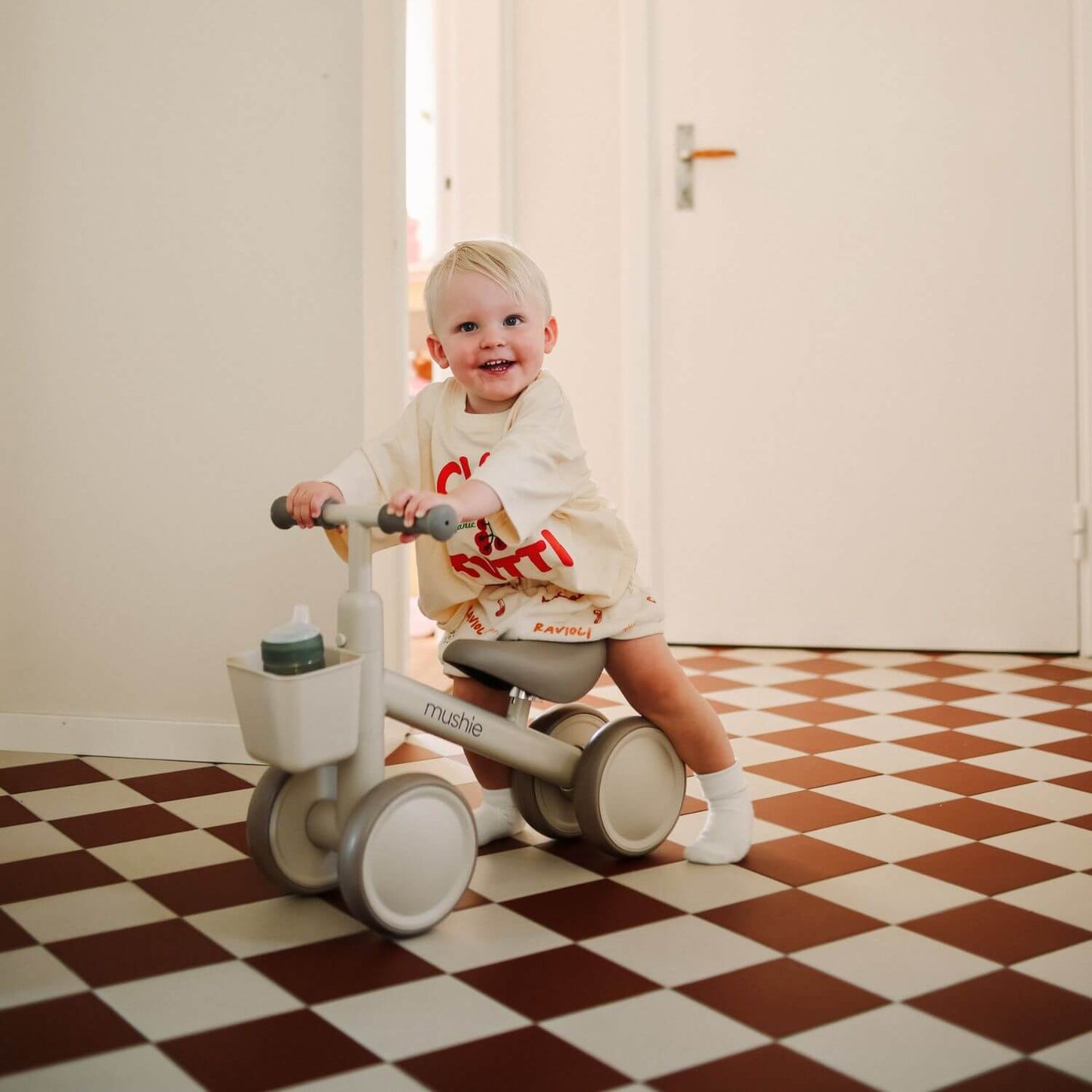 A toddler sits on Mushie Mini Toddler Bike in a room with a brown-and-white checkered floor and cream walls.