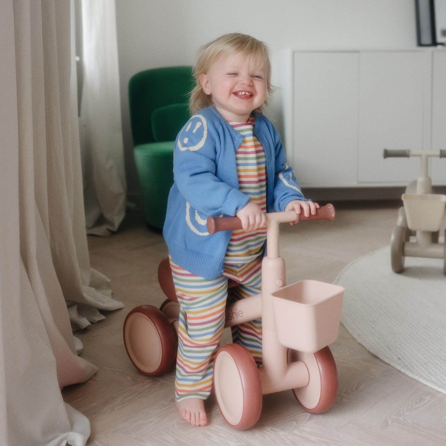 Child in a colorful outfit standing next to Mushie Mini Toddler Bike in a room with light-colored walls and furniture.