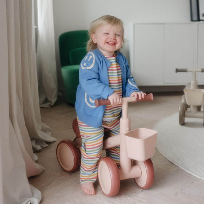 Child in a colorful outfit standing next to Mushie Mini Toddler Bike in a room with light-colored walls and furniture.