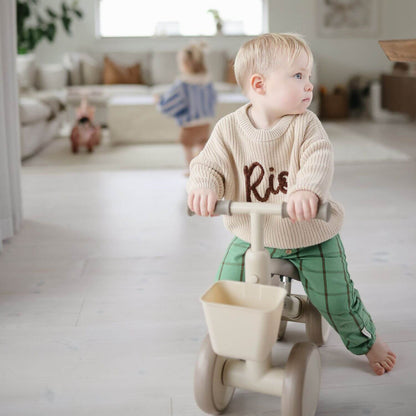 A toddler sits on Mushie Mini Toddler Bike in a bright, modern living room