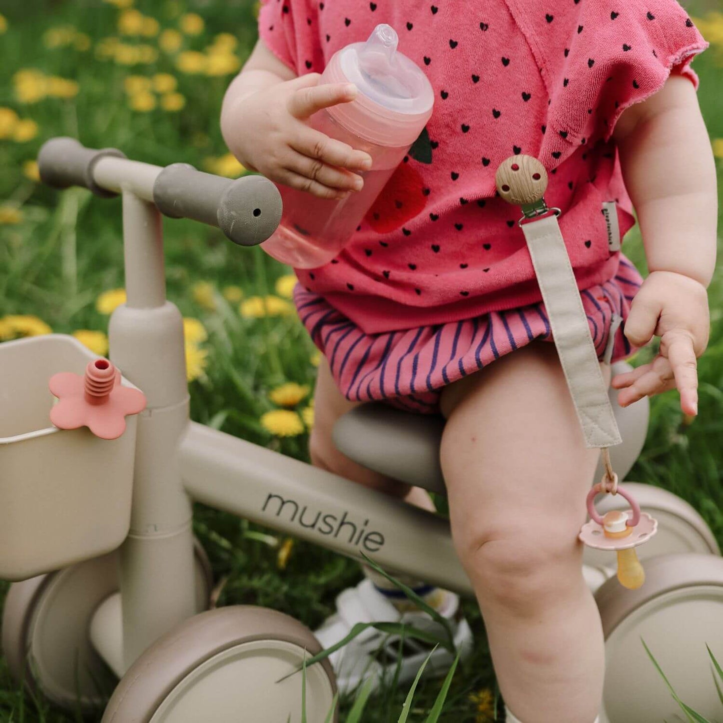Child on Mushie Mini Toddler Bike with a sippy cup and pacifier, sitting in a grassy area.