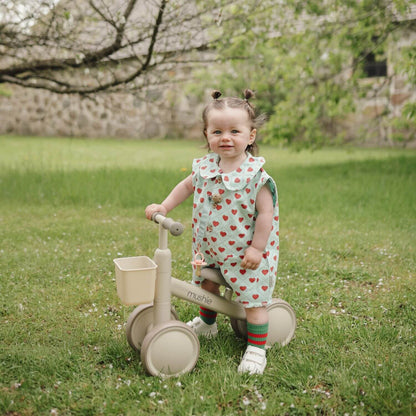Child in a heart-patterned dress riding Mushie Mini Toddler Bike in a grassy area with cherry blossoms.