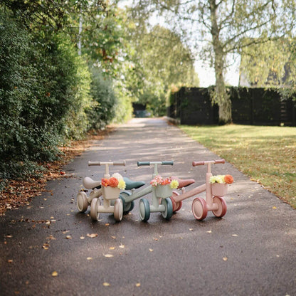 Three Mushie Mini Toddler Bikes on a paved path with greenery in the background