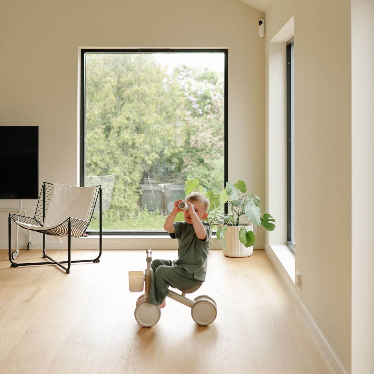 Child playing with Mushie Mini Toddler Bike in a modern living room with large windows.