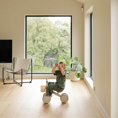 Child playing with Mushie Mini Toddler Bike in a modern living room with large windows.