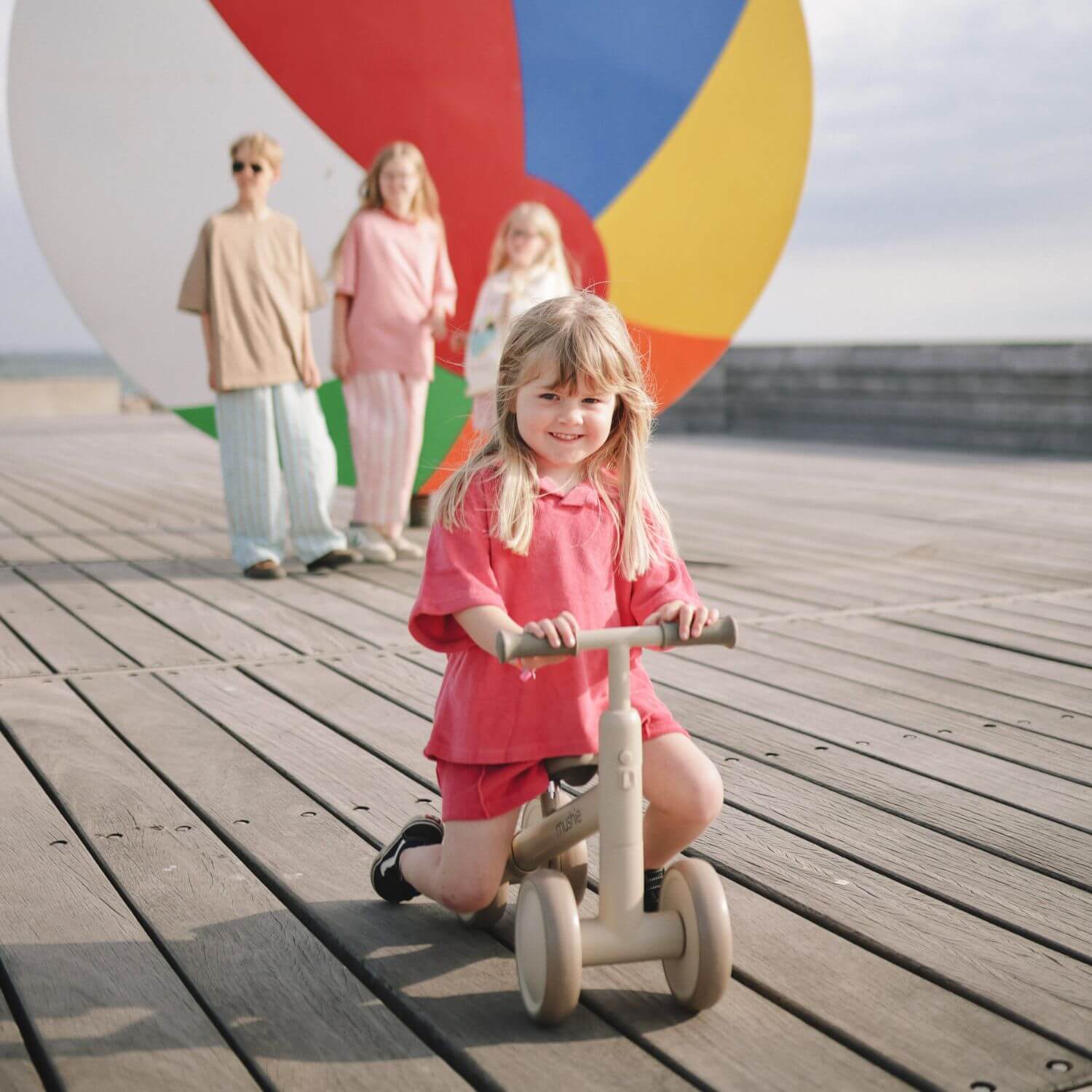 Child on Mushie Mini Toddler Bike with family in the background at a beach.