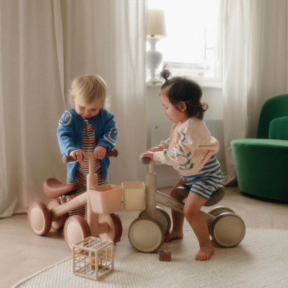 Two children playing with Mushie Mini Toddler Bikes in a room with curtains and a green chair.