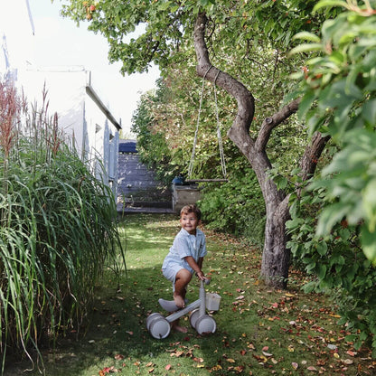 Child playing with Mushie Mini Toddler Bike in a garden with trees and plants.