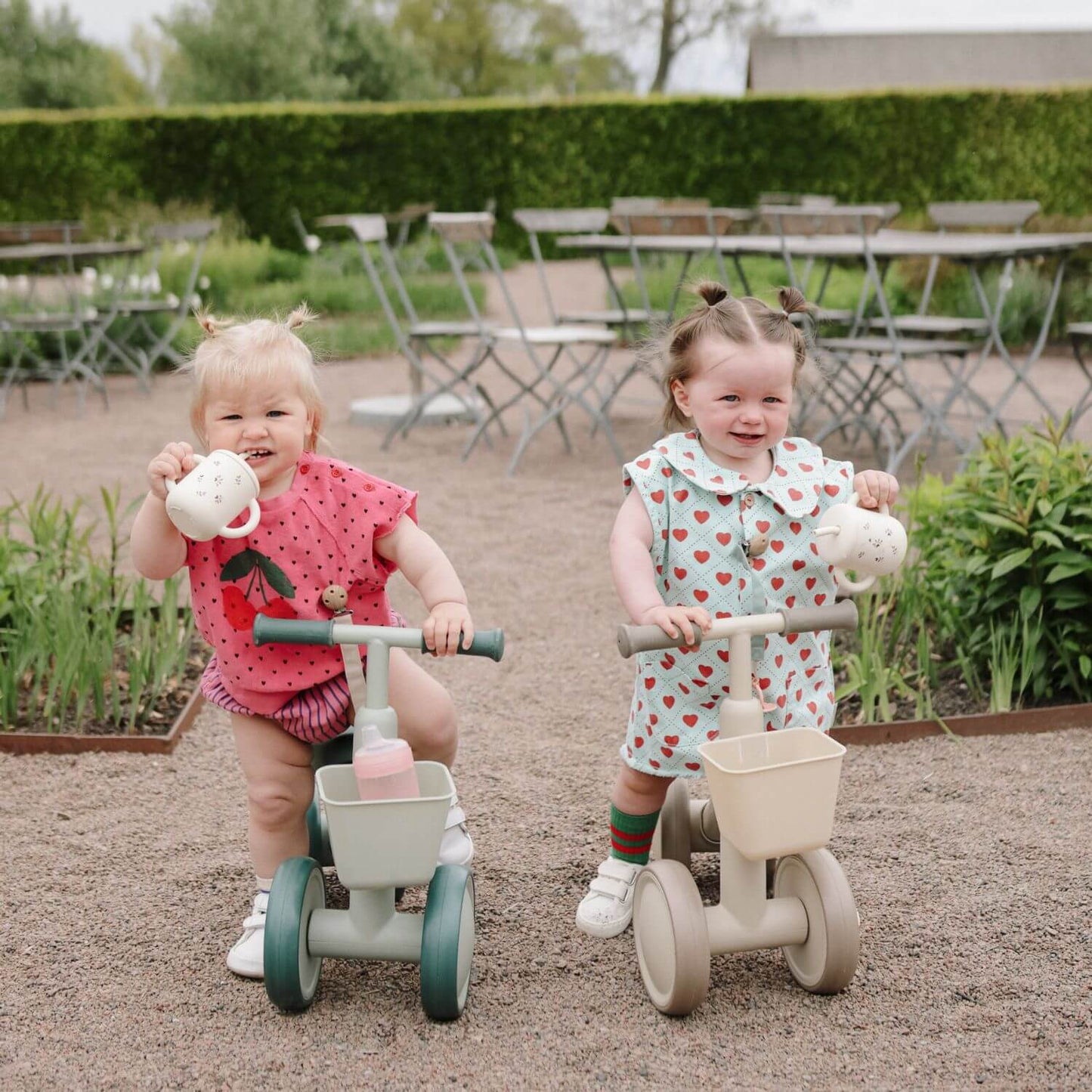 Two young children on Mushie Mini Toddler Bikes in an outdoor setting with tables and chairs in the background.