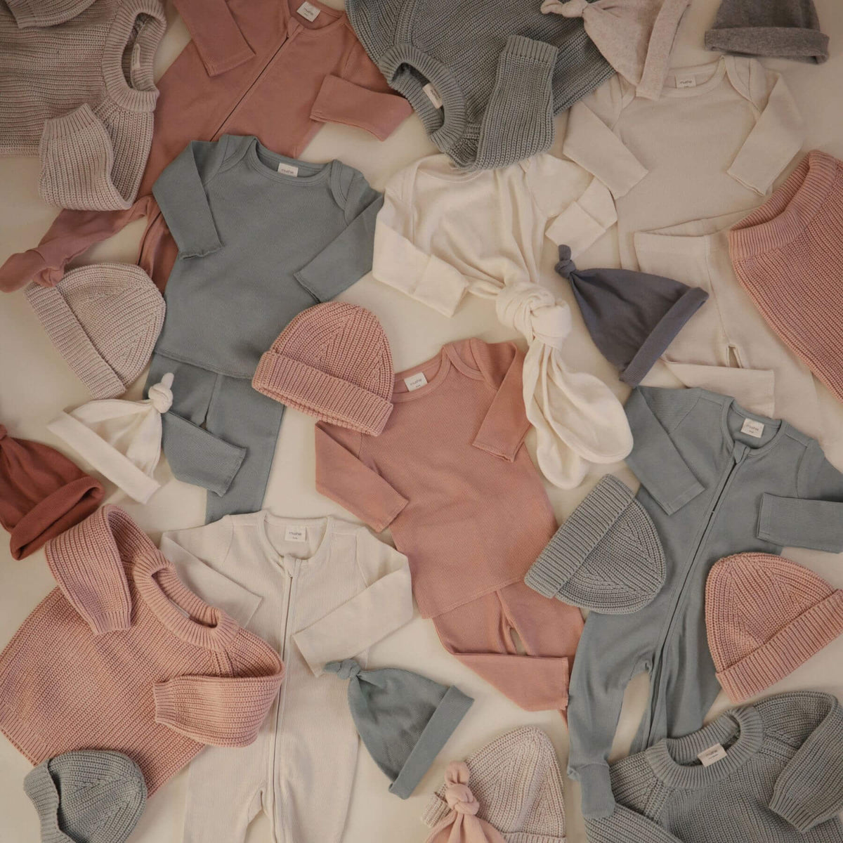 Collection of Mushie baby clothes including sweaters and hats in various colors on a light background