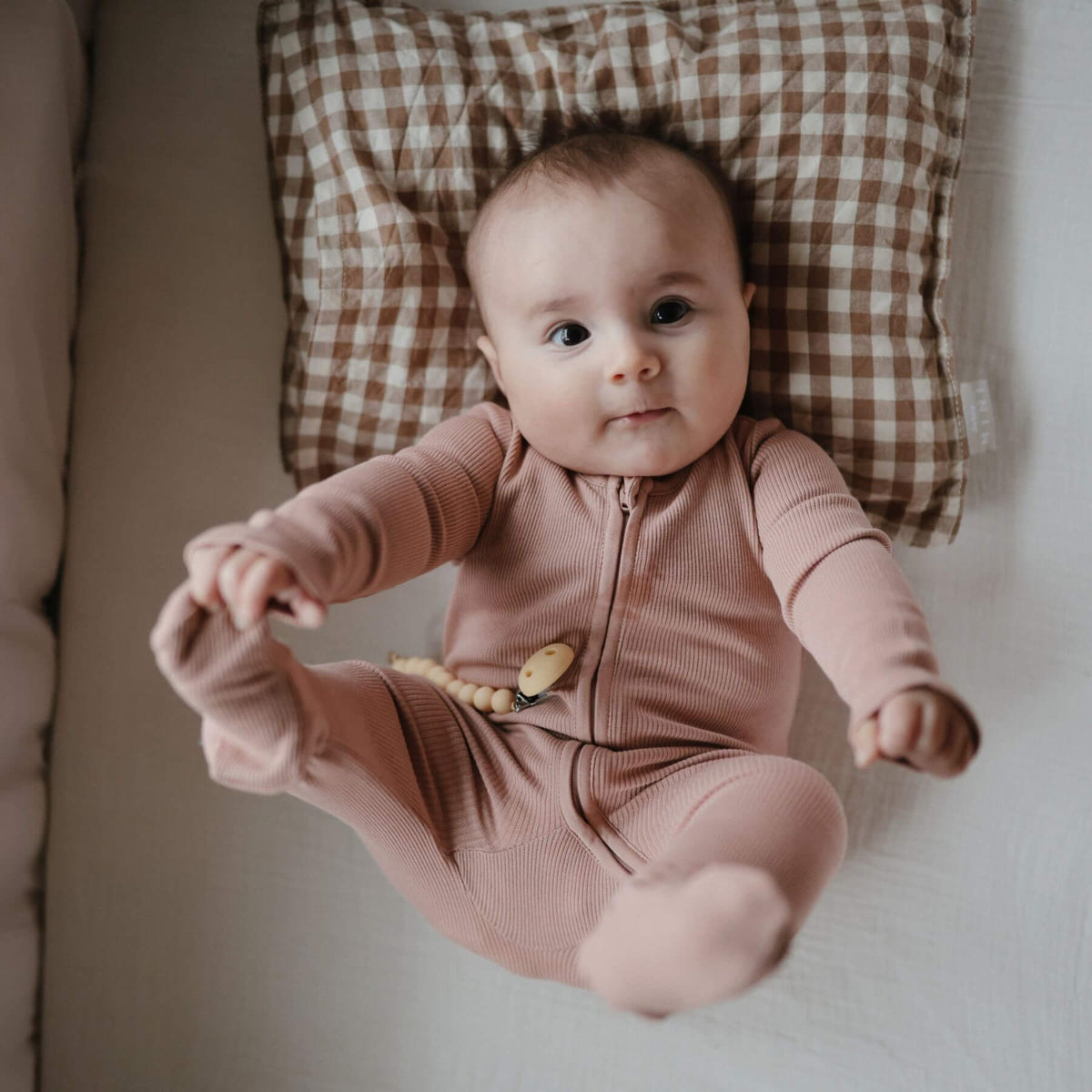 Baby in Mushie Zipper Pajama - Blush lying on a checkered cushion