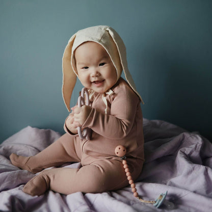 Baby in Mushie Zipper Pajama - Blush sitting on a purple blanket against a teal background